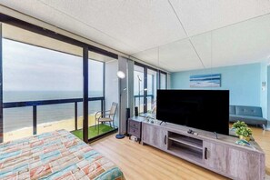 A bedroom with a large bed, a TV, and a balcony with a view of the ocean. - Beachfront studio with ocean views, AC, W/D, balcony, pool, gym & beach access (Ocean City)