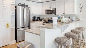 Una cocina moderna con armarios blancos, electrodomésticos de acero inoxidable, encimera de mármol y taburetes de fibra natural.