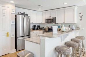 A modern kitchen with white cabinets, stainless steel appliances, a marble countertop, and woven stools. - 3rd-floor corner condo with amazing ocean views, balcony, rooftop pool, & W/D (Ocean City)