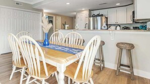 Un comedor con una mesa de madera y sillas blancas, una cocina con armarios blancos, y un taburete de bar.