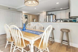 A dining area with a wooden table and white chairs, a kitchen with white cabinets, and a bar stool. - 3rd-floor corner condo with amazing ocean views, balcony, rooftop pool, & W/D (Ocean City)