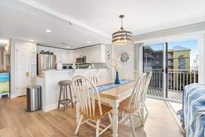 A modern kitchen with white cabinets, a stainless steel refrigerator, and a dining area with a wooden table and white chairs. - 3rd-floor corner condo with amazing ocean views, balcony, rooftop pool, & W/D (Ocean City)