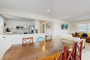 A modern kitchen with white cabinets, a dining table with wooden chairs, a living area with a sofa and cushions, and a television mounted on the wall. - Large two-level home with two kitchens, two decks, fast WiFi, & great location (Ocean City)