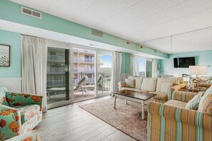 A living room with a sofa, armchair, and a coffee table. There is a sliding glass door leading to an outdoor area with a balcony. - Blue Sea Colony 202 (Ocean City)