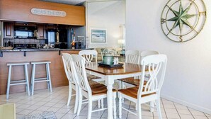 A dining area with a wooden table and white chairs, a kitchen area with a sign reading 'SEAS THE DAY,' and a compass wall decoration. - Beach-View Condo at the Pyramid - Shared Oceanfront Pool, WiFi & Central AC (Ocean City)