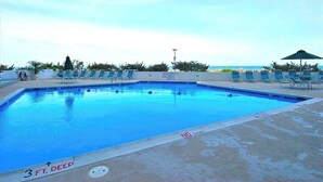A swimming pool with lounge chairs and umbrellas, a clear sky, and a view of the ocean. - Beach-View Condo at the Pyramid - Shared Oceanfront Pool, WiFi & Central AC (Ocean City)