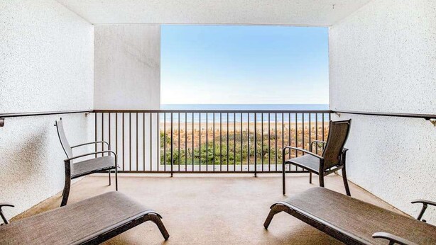 A balcony with two wooden chairs and a table, overlooking a beach and ocean. - Beach-View Condo at the Pyramid - Shared Oceanfront Pool, WiFi & Central AC (Ocean City)