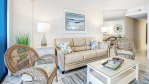 A living room with a beige sofa, wicker chairs, a glass coffee table, and a wall-mounted painting. - Beach-View Condo at the Pyramid - Shared Oceanfront Pool, WiFi & Central AC (Ocean City)