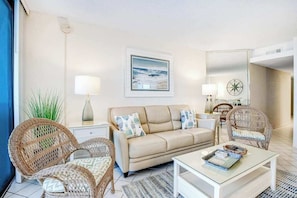 A living room with a beige sofa, wicker chairs, a glass coffee table, and a wall-mounted painting. - Beach-View Condo at the Pyramid - Shared Oceanfront Pool, WiFi & Central AC (Ocean City)