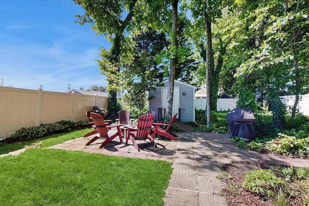 Un jardin avec une terrasse, des fauteuils Adirondack rouges, un barbecue et un abri de jardin.