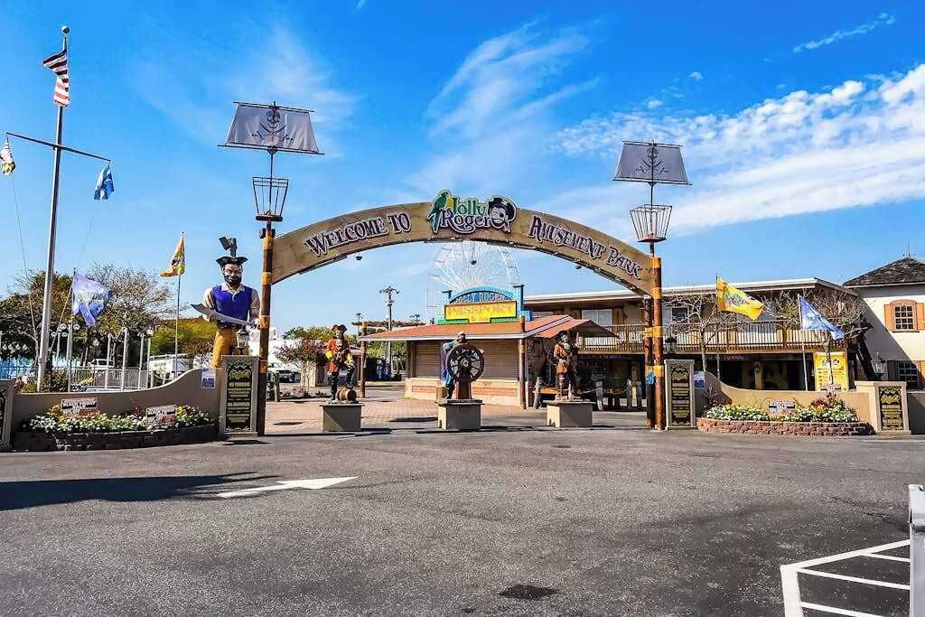 Un arco de bienvenida con el letrero 'Jolly Roger Amusement Park', una noria y un edificio con un cartel.