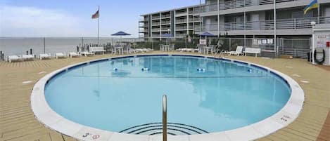 A pool area with lounge chairs and umbrellas, a flag, and a building with balconies.