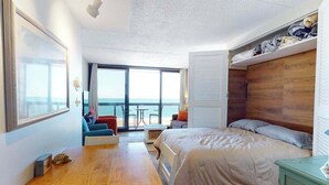 A bedroom with a bed, a wooden headboard, a dining table, a sofa, and a view of the ocean. - Delightful beachfront studio with full kitchen, balcony, pools, gym & tennis (Ocean City)