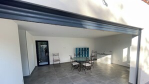 A spacious indoor area with a dining table and chairs, a kitchen area, and a door leading to another room. - Villa des amis (Caissargues)