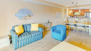 Ein Wohnzimmer mit einer Couch, einem Sessel, einem Esstisch mit Stühlen, einem kleinen Beistelltisch und einer Küchenzeile mit Schränken und einem Kühlschrank.