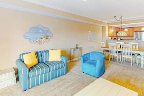 A living room with a sofa, a chair, a dining table with chairs, a small side table, and a kitchen area with cabinets and a refrigerator. - Coral Seas 103 (Ocean City)