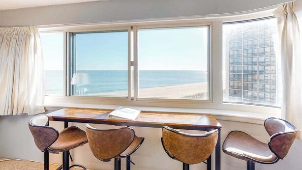 Quarto com uma grande janela em arco com vista para o mar, uma mesa de madeira e quatro cadeiras.