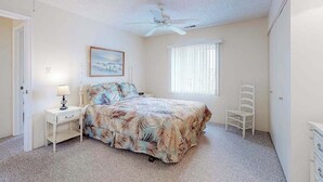 Une chambre à coucher comprenant un lit, une table de chevet, une chaise, un ventilateur de plafond, une fenêtre avec des stores et un tableau accroché au mur.