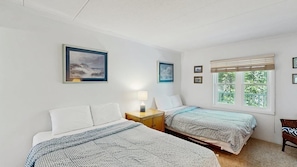 Et soveværelse med to senge, et natbord med lampe, et vindue med skodder, og billeder i rammer på væggen.