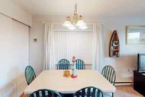 Ein Essbereich mit Tisch, Stühlen, einer Deckenleuchte, Vorhängen, einem Fernseher und einem Regal mit Gegenständen.