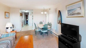 A living room with a sofa, a dining table with chairs, a TV, and a kitchen in the background. - Partial Ocean View Condo in Oceanfront Building with Balcony, WiFi & Laundry (Ocean City)