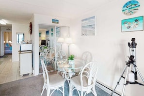 A dining area with a glass table and white chairs, a lamp, and a telescope on a tripod. - Oceanfront Condo with Direct Beach View, Balcony, AC, WiFi & Laundry (Ocean City)