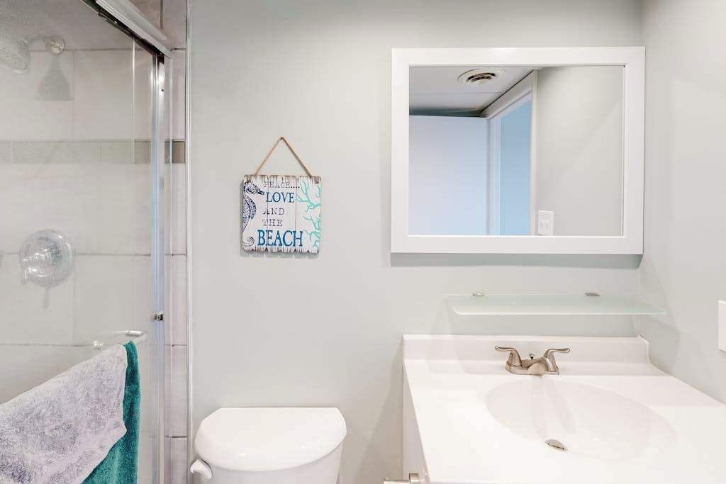 Ein Badezimmer mit Duschkabine aus Glas, einer weißen Toilette, einem weißen Waschbecken mit Spiegel darüber und einem an der Wand befestigten Schild mit der Aufschrift 'LOVE AND THE BEACH'.