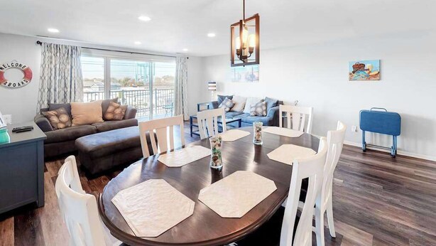 A modern living room with a dining area, a sofa set, a blue suitcase, and a cityscape painting. - Condo One Block from the Beach w/ Balcony, AC, Laundry & Shared Pool (Ocean City)