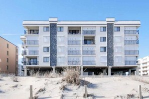 A multi-story building with balconies, a flat roof, and a sandy ground in front. - Waikiki 302 (Ocean City)