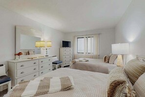 Ein Hotelzimmer mit zwei Betten, einem Schminktisch, einem Fernseher und einem Fenster mit Vorhängen.