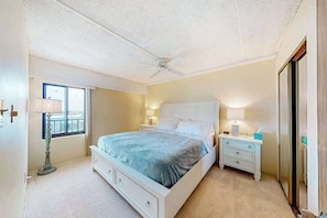 A bedroom with a bed, bedside tables, a ceiling fan, a window, and a door. - 7th-floor condo with sunset & ocean views, pool, balcony, outdoor shower, W/D (Ocean City)