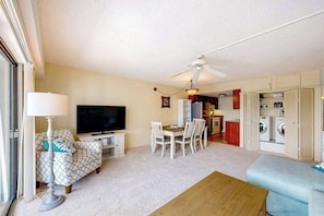 A living room with a television, dining table, chairs, a sofa, a coffee table, a ceiling fan, and a laundry area with washing machines. - 7th-floor condo with sunset & ocean views, pool, balcony, outdoor shower, W/D (Ocean City)