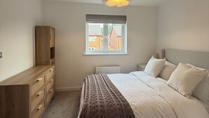 A bedroom with a bed, a wooden dresser, a window with a view, and a hanging light fixture. - New Home w/2+ car parking and charging spaces (Knutsford)