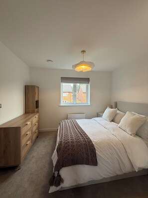 A bedroom with a bed, a wooden dresser, a window with a view, and a hanging light fixture. - New Home w/2+ car parking and charging spaces (Knutsford)