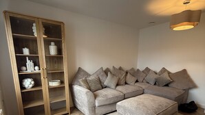 A modern living room with a herringbone wood floor, a grey sectional sofa with multiple cushions, a wooden cabinet with glass doors, and two pendant lights. - New Home w/2+ car parking and charging spaces (Knutsford)