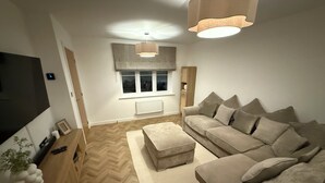 A living room with a sofa, ottoman, TV, wooden side table, and a large window with curtains. - New Home w/2+ car parking and charging spaces (Knutsford)