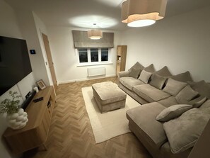 A living room with a sofa, ottoman, TV, wooden side table, and a large window with curtains. - New Home w/2+ car parking and charging spaces (Knutsford)