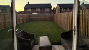 A patio with wicker furniture, a small table, and a view of a residential area with houses and a wooden fence. - New Home w/2+ car parking and charging spaces (Knutsford)