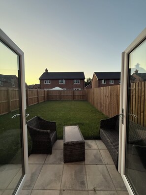 A patio with wicker furniture, a small table, and a view of a residential area with houses and a wooden fence. - New Home w/2+ car parking and charging spaces (Knutsford)