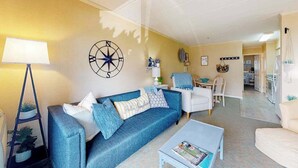 A living room with a sofa, coffee table, and a lamp. There is a kitchen in the background. - Ocean block condo in Ocean City with balcony & steps to the beach (Ocean City)