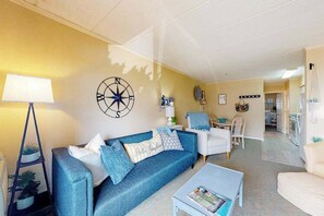 Ein Wohnzimmer mit Sofa, Couchtisch und Lampe. Im Hintergrund ist eine Küche zu sehen.