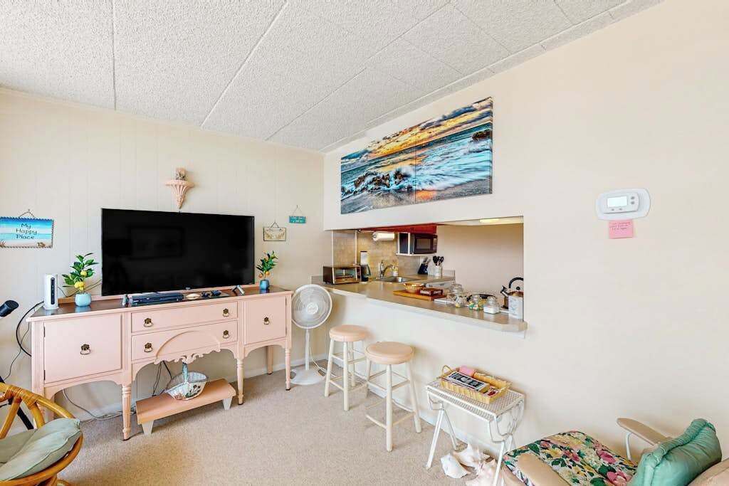 A living room with a TV, pink furniture, a kitchen area, and a painting on the wall.