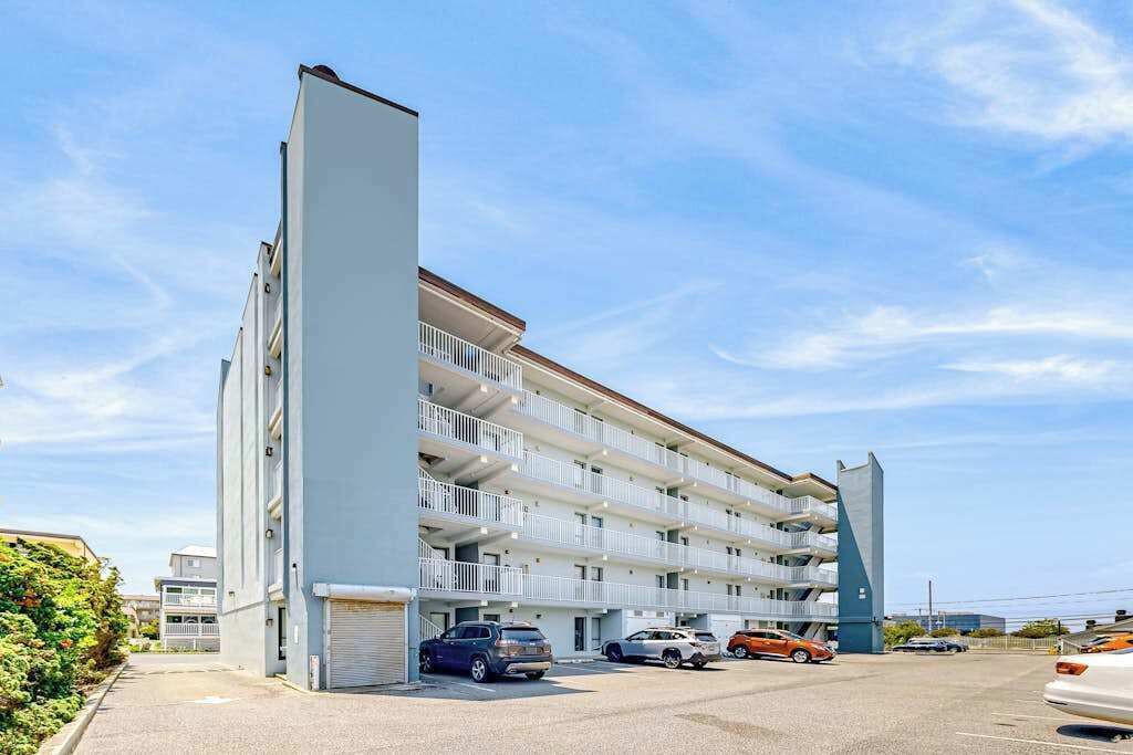 A multi-story building with balconies, a parking area, and a clear sky.
