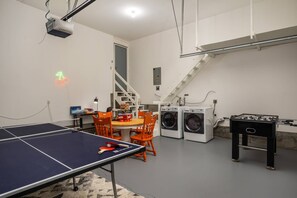 A room with a ping pong table, two chairs, a washing machine, a foosball table, and a staircase.