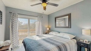 Un dormitorio con cama, ventilador de techo, ventana con cortinas, mesita de noche con lámpara y espejo en la pared.