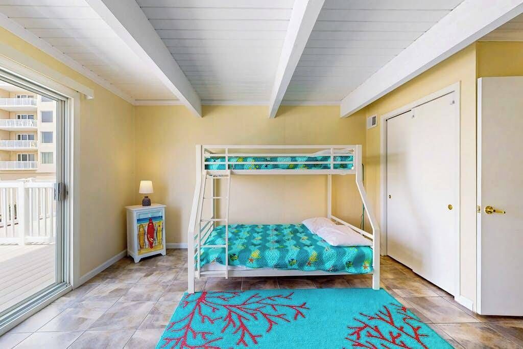 Habitación con literas y vistas a un balcón, una mesita de noche con lámpara, y una puerta a la derecha.