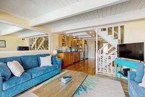 Ein geräumiger Wohnbereich mit einer Couch, einem Couchtisch und einem Fernseher. Im Hintergrund ist eine Küche erkennbar, und eine Treppe führt zu einer weiteren Ebene.