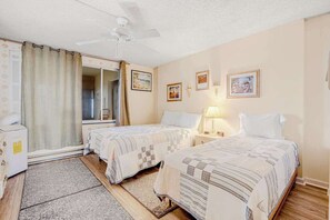Ein Hotelzimmer mit zwei Betten, einem Deckenventilator, einem Fenster mit Vorhängen, einem Nachttisch mit Lampe und gerahmten Bildern an der Wand.