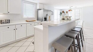A modern kitchen with white cabinets, a stainless steel refrigerator, a built-in island with bar stools, and a window with blinds. - Family-Friendly Beach Condo: Balcony, w/d, shared outdoor pool & gym (Ocean City)