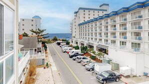 A coastal hotel with multiple-story buildings, a parking lot with cars, and a view of the ocean. - Family-Friendly Beach Condo: Balcony, w/d, shared outdoor pool & gym (Ocean City)